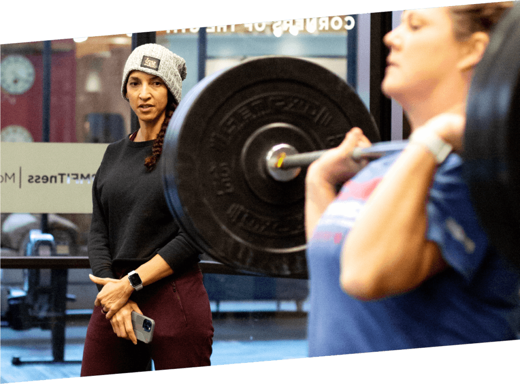 Nicole, CPM coach, watches attentively as a gym member holds a barbell in a front rack position.